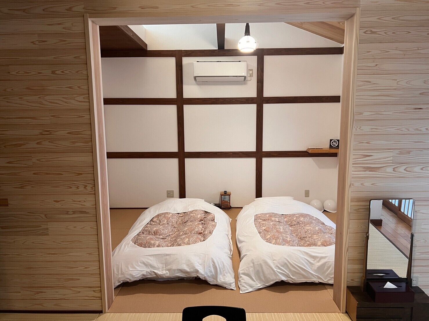 A room with a traditional Japanese-style interior, featuring tatami flooring, a low ceiling, and a sliding door leading to a bed with two pillows.