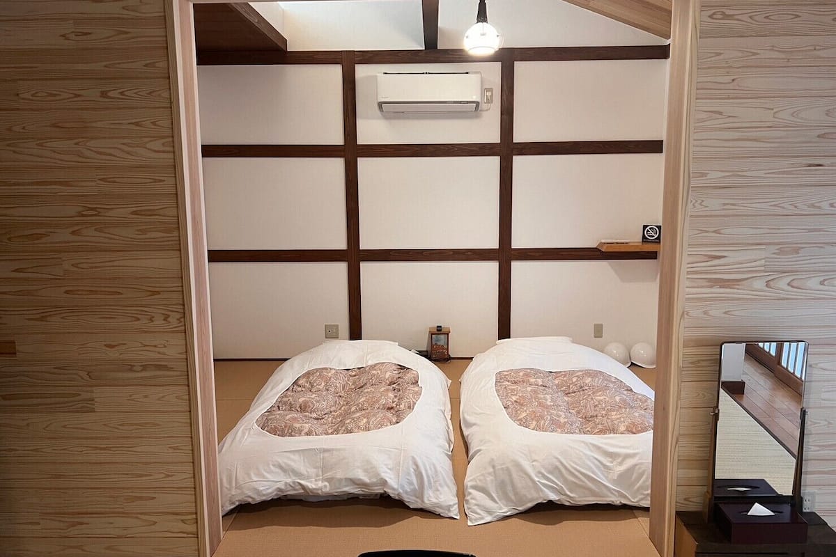 A room with a traditional Japanese-style interior, featuring tatami flooring, a low ceiling, and a sliding door leading to a bed with two pillows.