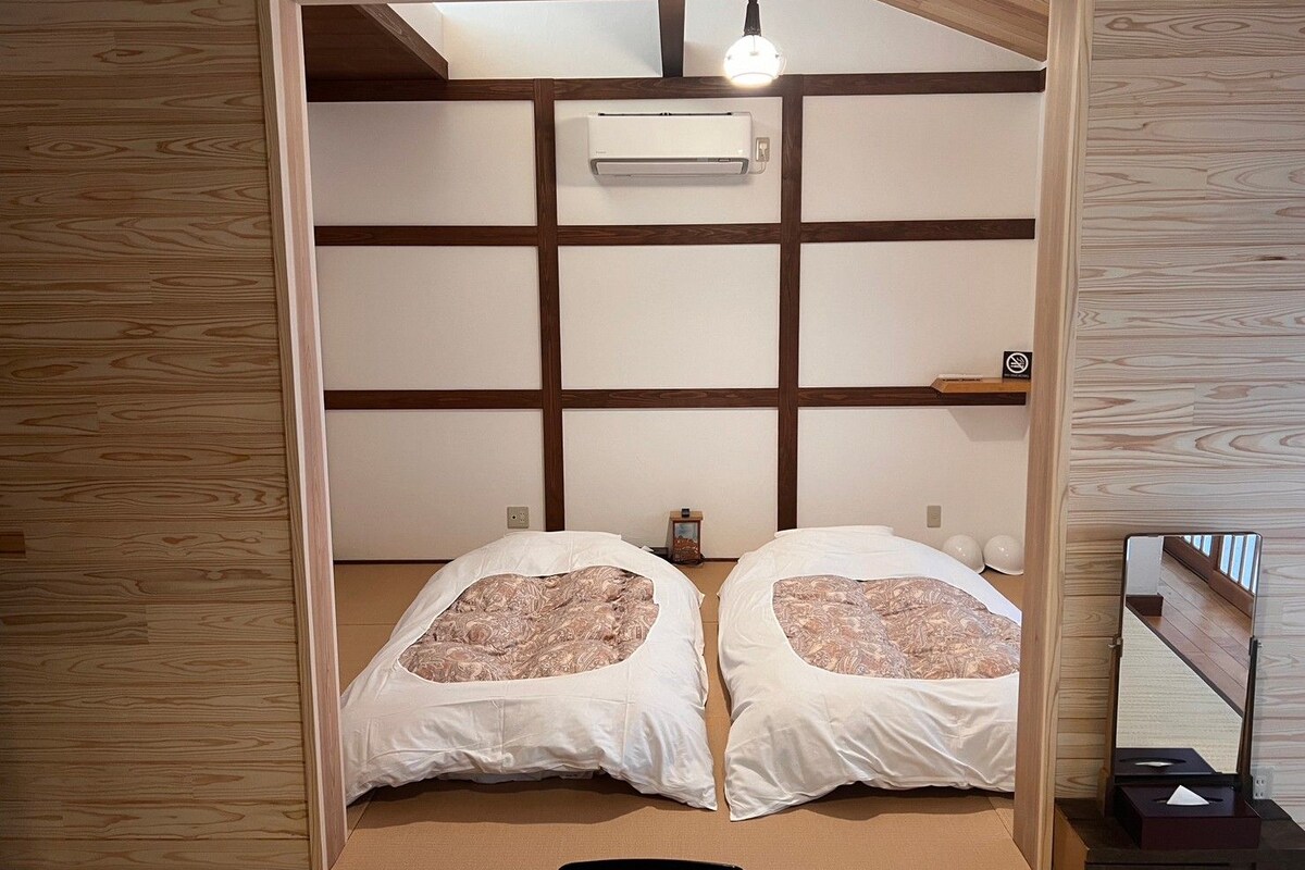 A room with a traditional Japanese-style interior, featuring tatami flooring, a low ceiling, and a sliding door leading to a bed with two pillows.