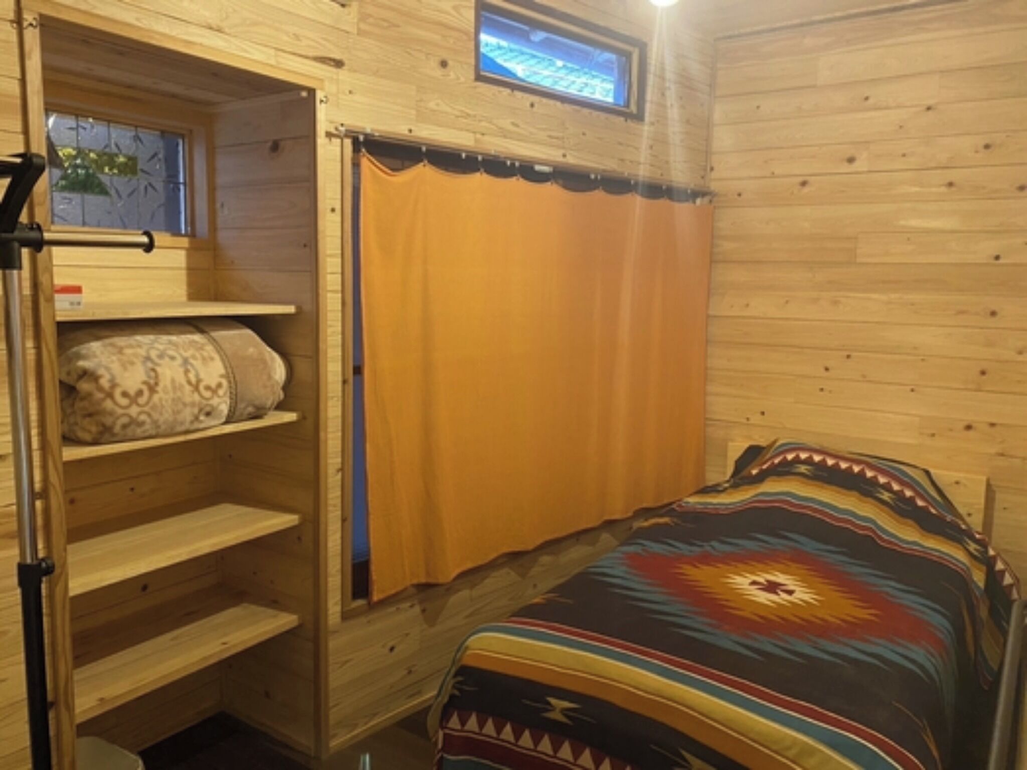 A wooden cabin room with a bed, a window with curtains, and a shelf with pillows.