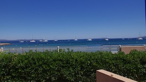 Vista costera con cielo despejado, un puerto deportivo con varios veleros y un seto en primer plano.