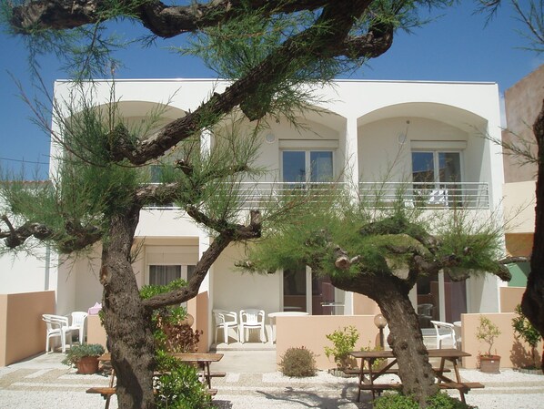 Un edificio con fachada blanca, varias ventanas y un balcón. Hay mesas y sillas al aire libre, y un árbol en primer plano.