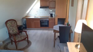 A compact kitchen with wooden cabinets, a built-in oven, and a sink. There is a dining table with a chair, a lamp, and a remote control on a wooden surface.