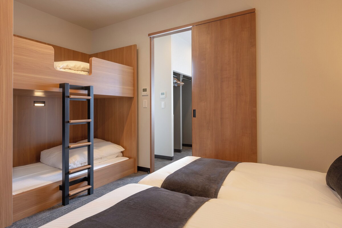 A hotel room with a bunk bed, a separate bed, a wardrobe, and a bathroom visible through an open door.