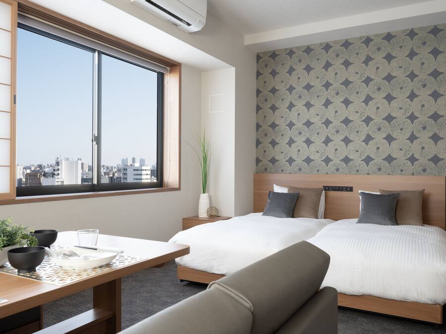 Habitación de hotel con dos camas, una mesa de comedor, una planta en maceta y vistas a la ciudad.
