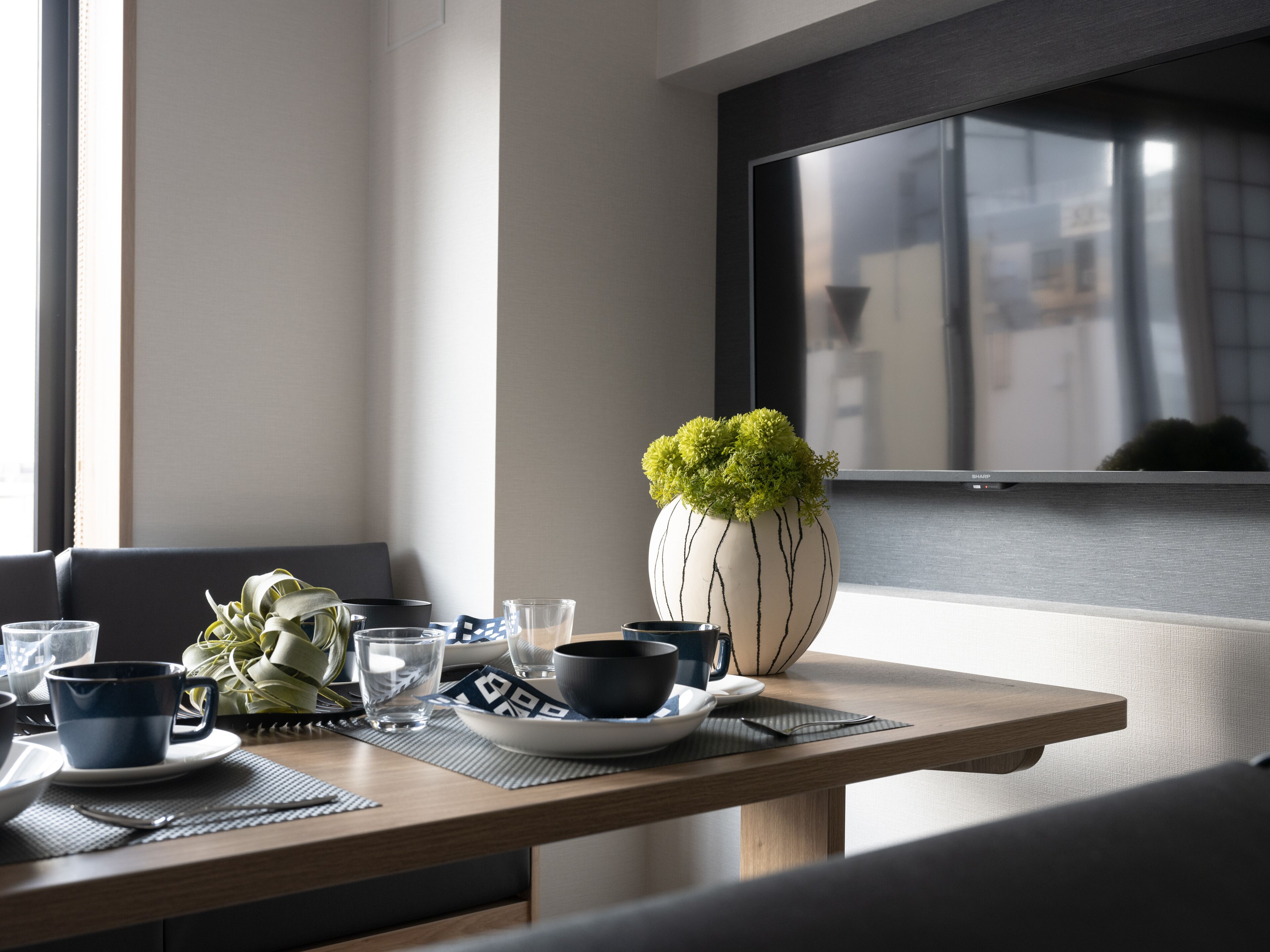Un coin repas avec une table dressée pour deux personnes, agrémenté d’un vase contenant des feuilles vertes, d’un bol et d’une tasse.