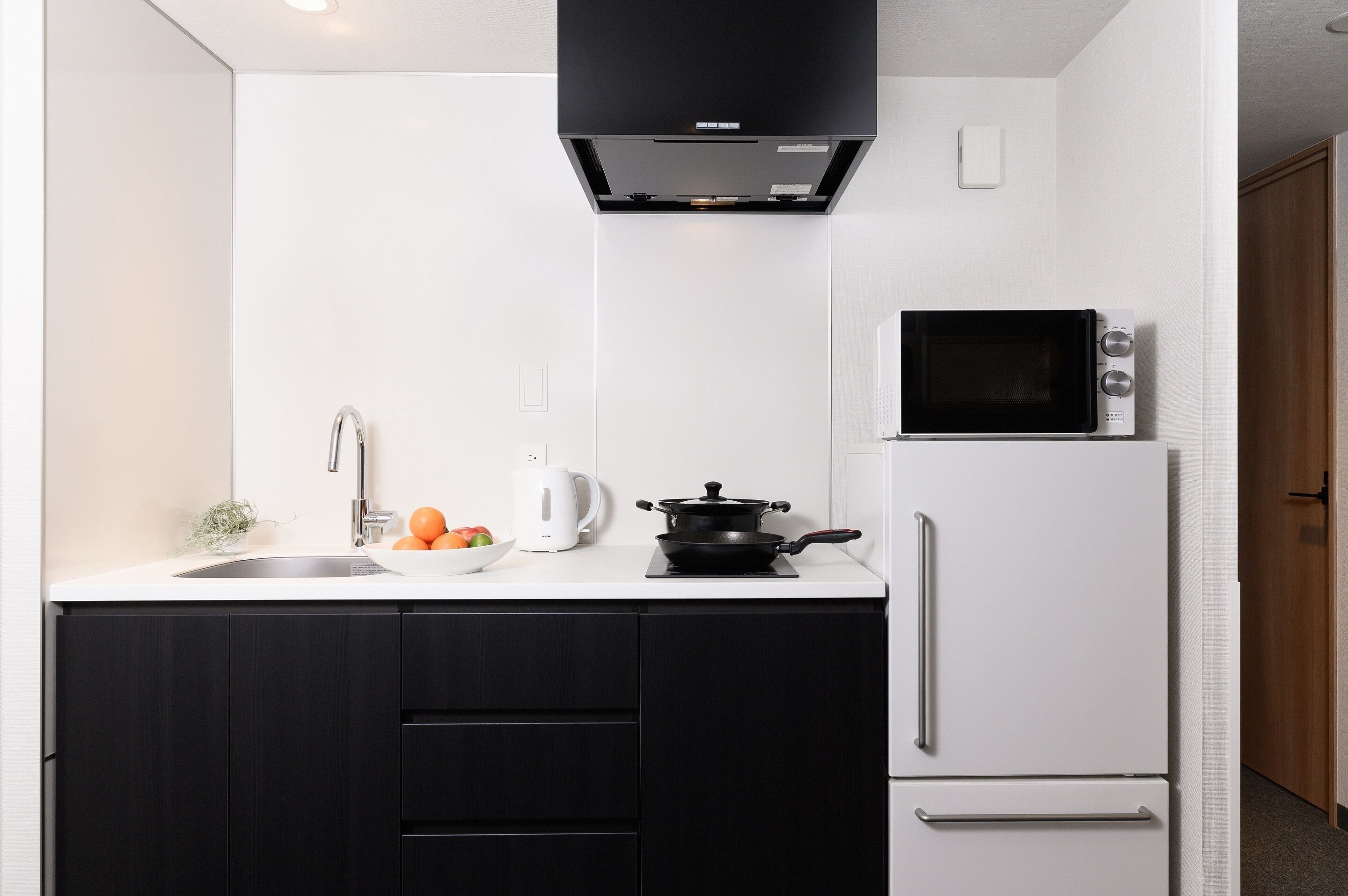A modern kitchen with a black range hood, a microwave, a white refrigerator, and a sink with a kettle and oranges on the counter.
