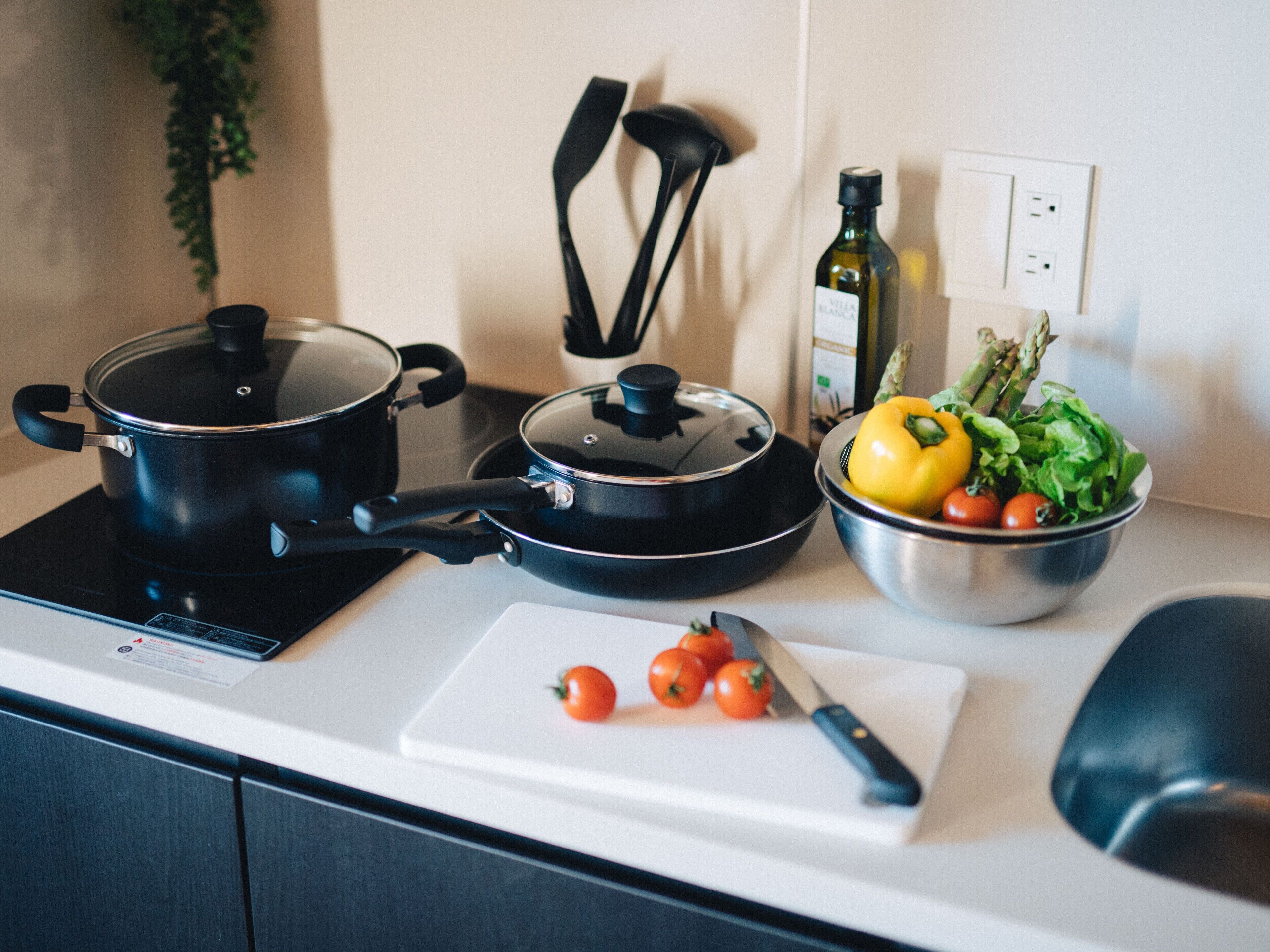 A kitchen with two black pots on an induction cooktop, a cutting board with tomatoes, a knife, a bowl of vegetables, and a bottle of olive oil on the counter.