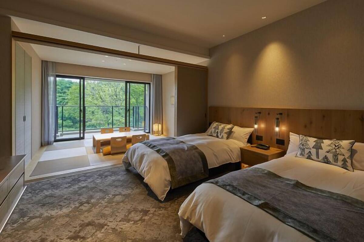 Quarto de hotel moderno com duas camas, área de refeições e varanda com vista para o verde.