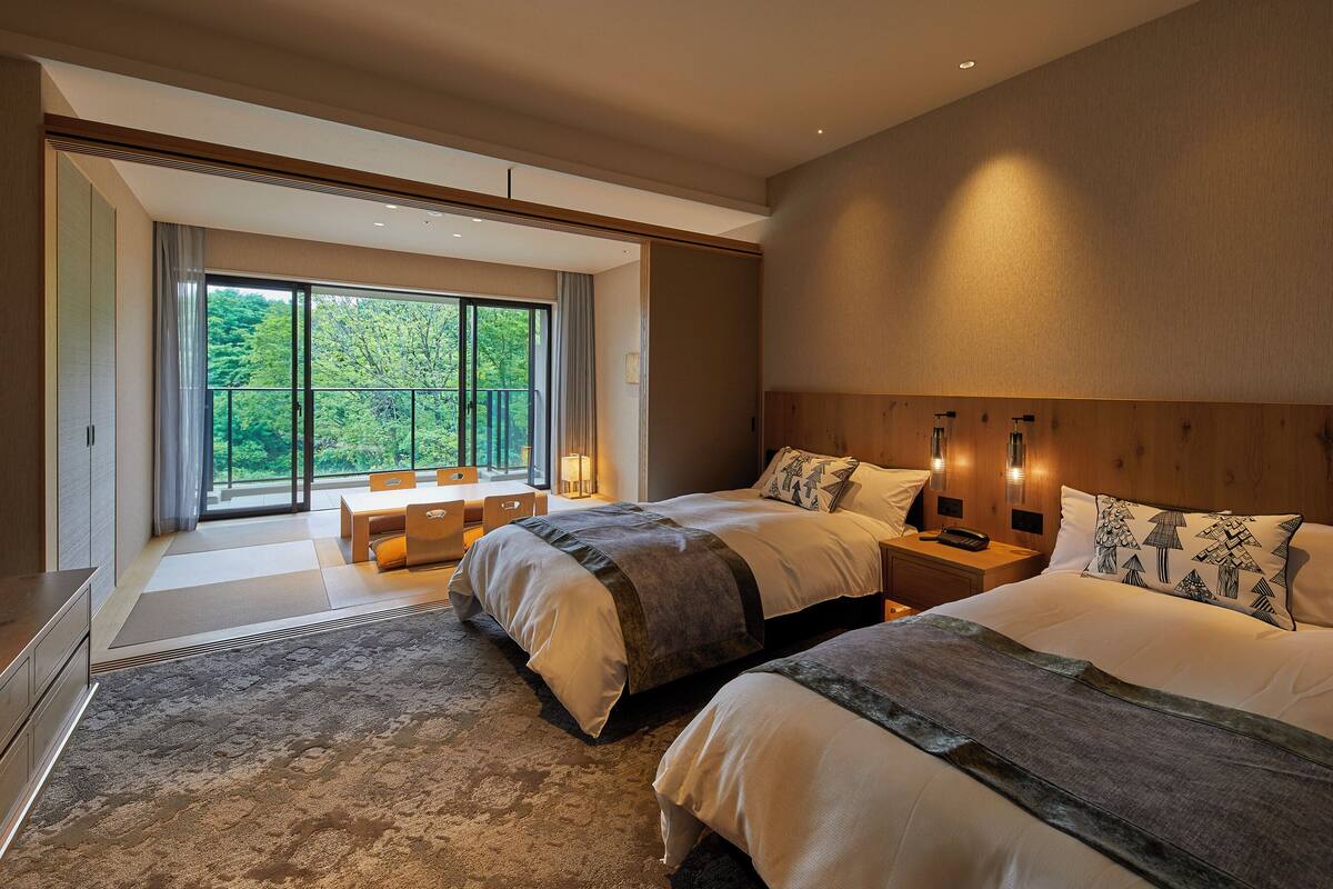 Quarto de hotel moderno com duas camas, área de jantar e varanda com vista para áreas verdes.