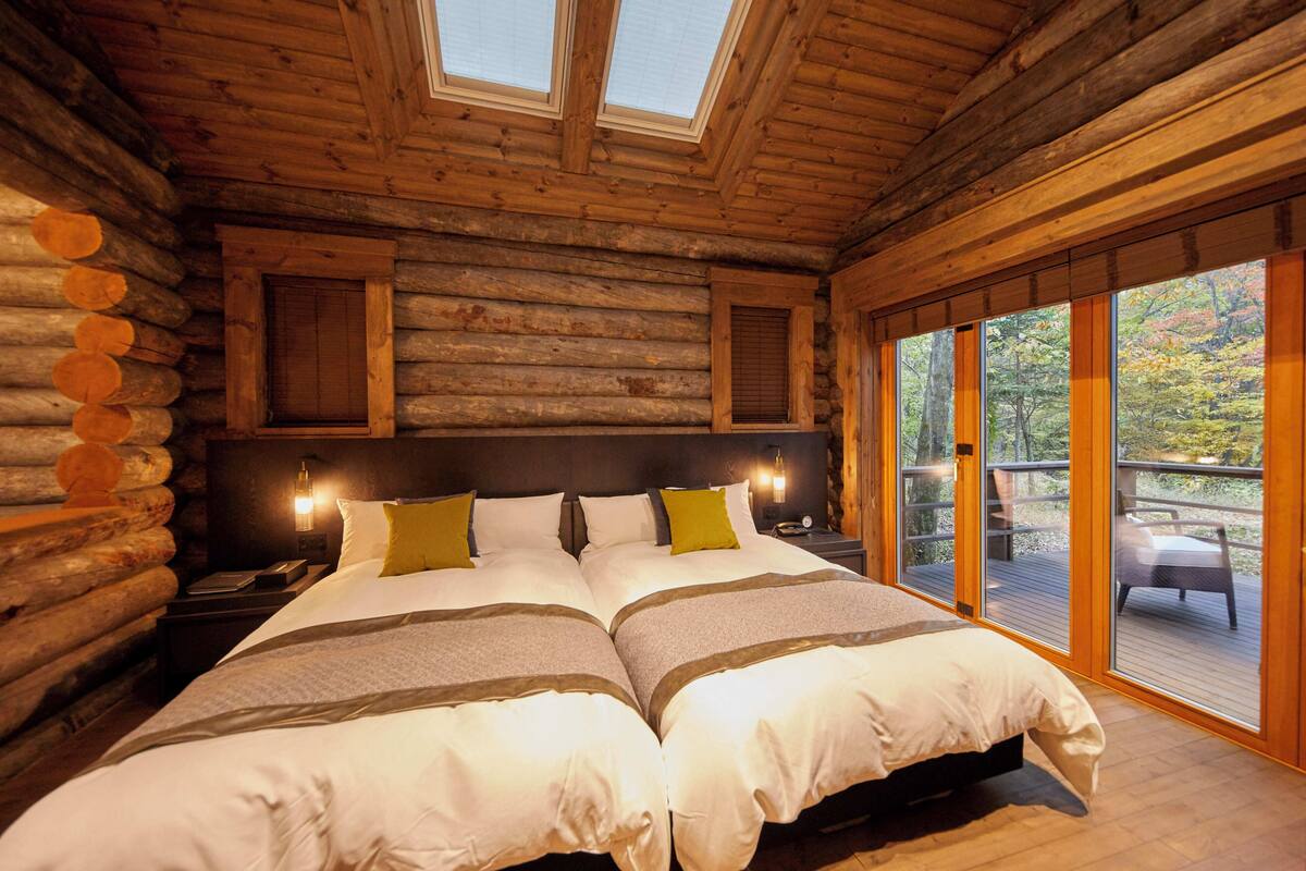 Quarto de cabana de madeira com duas camas, paredes rústicas, claraboia e varanda com vista para árvores.