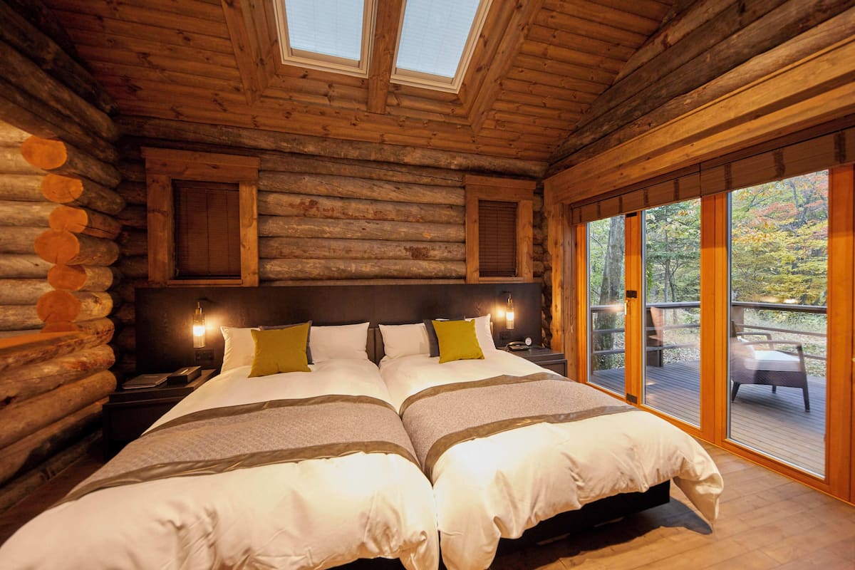 A log cabin bedroom with two beds, wooden walls, a skylight, and a balcony with a view of trees.