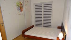 A bedroom with a wooden bed, a nightstand with a lamp, a wardrobe with a tree decal, and a window with blinds. - New 50m² apartment in Bertrange with basement parking (Bertrange)