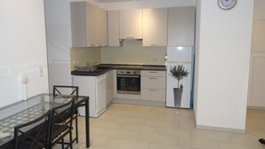 A compact kitchen with white cabinets, a dishwasher, an oven, and a refrigerator. There is a dining table with chairs and a small potted plant on the counter. - New 50m² apartment in Bertrange with basement parking (Bertrange)