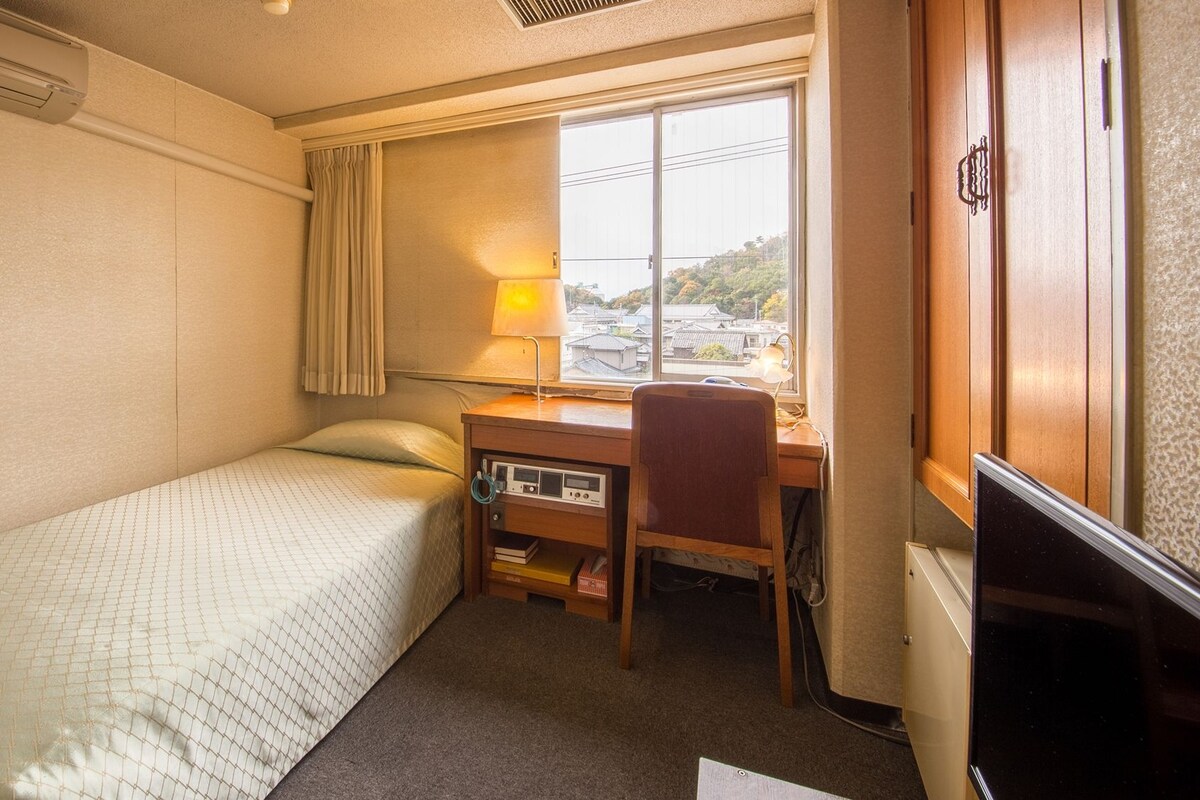 A hotel room with a single bed, a desk with a chair, a TV, and a window with curtains.