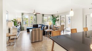 Een ruime woonkamer met een bank, eettafel en stoelen. Er zijn grote ramen met gordijnen, een plafondventilator en een plant.