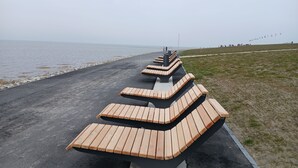A series of wooden benches arranged in a curved formation along a paved path by the water. - First floor vacation apartment Roseneck on the North Sea in Schleswig-Holstein, (Friedrichskoog)