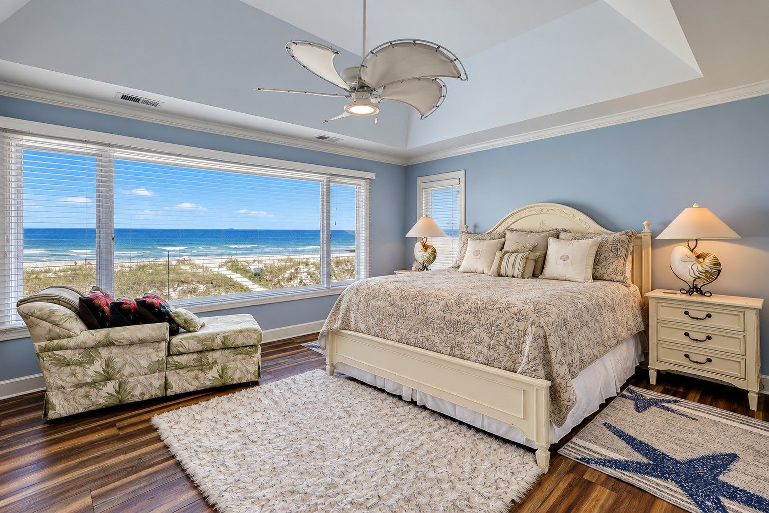 A bedroom with a large bed, a ceiling fan, a nightstand with a lamp, and a view of the beach.