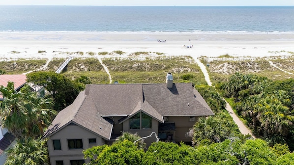 A house with a sloped roof, surrounded by lush greenery and a beach in the background. - 33 Sandpiper- Oceanfront, The NEW Daisy.  LARGE Private Pool & Fire Pit (Hilton Head Island)