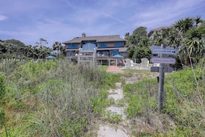 A two-story house with a pathway leading to the entrance, surrounded by tall grasses and palm trees. - 33 Sandpiper- Oceanfront, The NEW Daisy.  LARGE Private Pool & Fire Pit (Hilton Head Island)