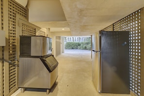 A corridor with a stainless steel refrigerator and a freezer, a concrete floor, and a view of a building with windows and a hedge in the distance. - 33 Sandpiper- Oceanfront, The NEW Daisy.  LARGE Private Pool & Fire Pit (Hilton Head Island)