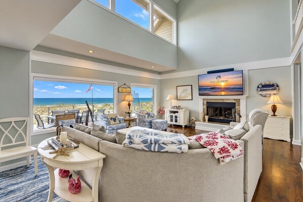 A spacious living room with a large window overlooking the beach, a fireplace, and a television mounted on the wall.