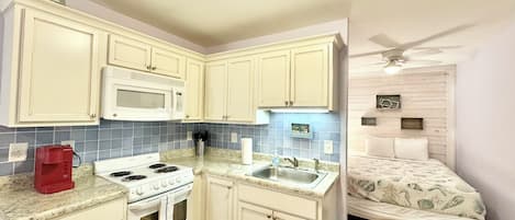 A compact kitchen with white cabinets, a microwave, a stove, and a sink. There's a small trash bin and a ceiling fan in the adjacent bedroom.
