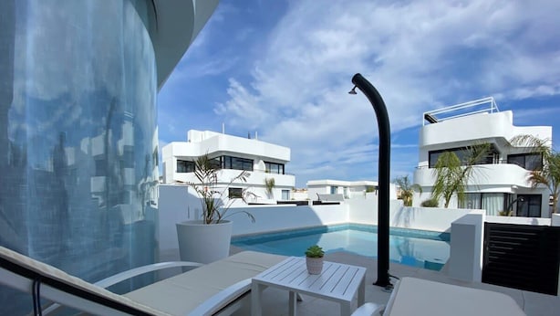 Un área de piscina moderna en la azotea, con edificios blancos, cielo despejado y un espacio de descanso junto a la piscina.