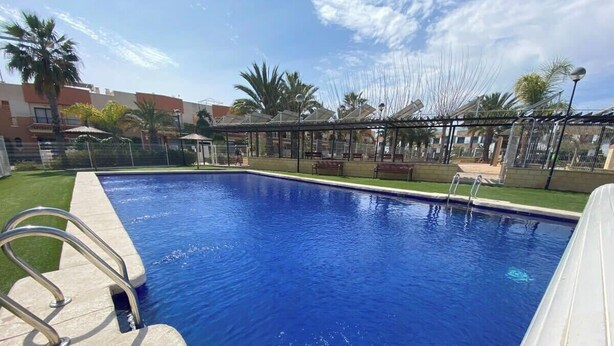 Ein rechteckiger Swimmingpool mit klarem, blauem Wasser, umgeben von einem Metallzaun und einer überdachten Fläche mit Bänken.