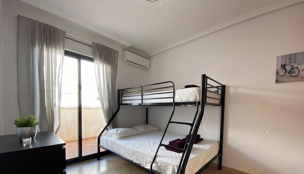 Habitación con literas, puerta corrediza al balcón, una mesita con una planta y aire acondicionado instalado en la pared.