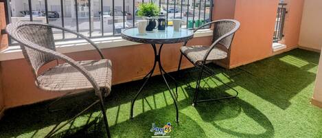 Un balcón con dos sillas de mimbre, una mesa de vidrio y una planta en maceta.