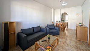 Un salon comprenant un canapé, un fauteuil et une table basse, un coin repas avec des chaises et une table, et une cuisine visible à travers une arche.