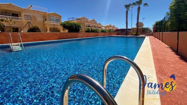 A swimming pool with clear blue water, a metal ladder, and a red brick walkway. - 2-Bed Ground-Floor Apt in La Cinuelica R2 L188 (Orihuela)