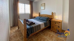 A bedroom with a wooden bed, a nightstand with a lamp, a window with curtains, and a framed picture on the wall.