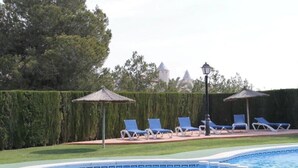 Un espace piscine agrémenté de chaises longues et de parasols, entouré d’une haie et d’arbres.