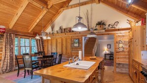Interior rústico de una cabaña de madera con una isla de cocina, zona de comedor y una escalera que sube a una buhardilla.