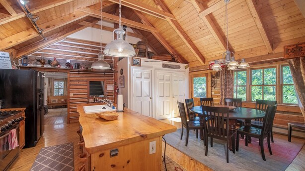 Interior rústico de una cabaña con una mesa de madera, sillas, una zona de cocina con estufa y refrigerador, y un ventanal que proporciona luz natural.