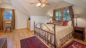 Un dormitorio con cama, ventilador de techo, ventana con cortinas, mesita de noche y una silla.