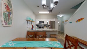 Een keuken met witte kastjes, een roestvrijstalen koelkast en een magnetron. Er staat een houten eettafel met stoelen en een blauwgroene placemat.