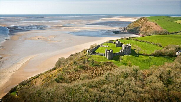 A coastal landscape with a castle on a grassy hill overlooking a beach and the sea.