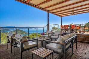 Een overdekt terras met houten meubels en uitzicht op zee.