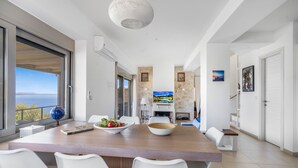 A modern dining area with a large table, white chairs, and a view of the sea.