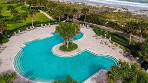 A pool with a curved design, surrounded by palm trees and lounge chairs, with a view of the ocean and beach in the background.