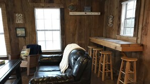 Un interior de cabaña acogedor, con un sofá de cuero, taburetes de madera y una ventana con persianas.