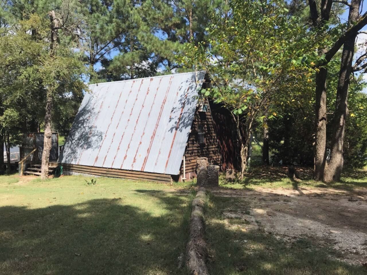 Una estructura de madera rústica con techo de lámina corrugada, rodeada de árboles y un área con césped.