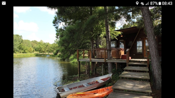 Una cabaña de madera con un muelle que se adentra en un lago tranquilo, rodeada de árboles.