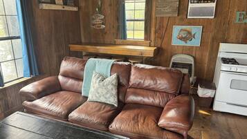 A rustic cabin interior with a leather couch, wooden walls, a window with blue curtains, a wooden shelf with fishing memorabilia, and a stove.