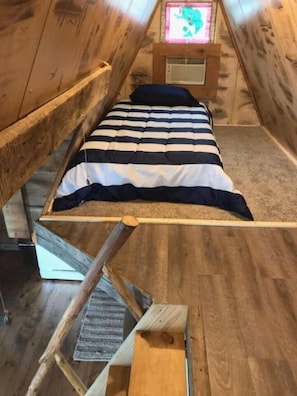 A loft-style bedroom with a bunk bed, wooden beams, a small window with a pink decoration, and a wooden ladder.
