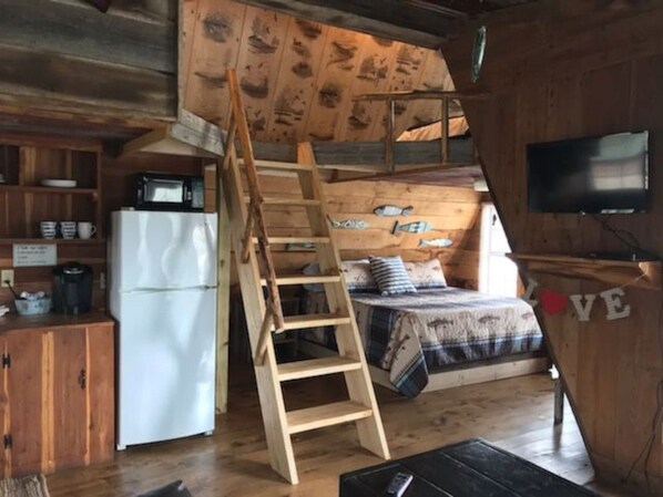 Een compact vakantiehuisje met een slaapzolder, een houten trap, een klein keukenhoekje en een aan de muur bevestigde televisie.