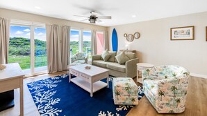 Un salon comprenant un canapé, une table basse et un pouf, un ventilateur de plafond et de grandes fenêtres donnant sur un paysage verdoyant.